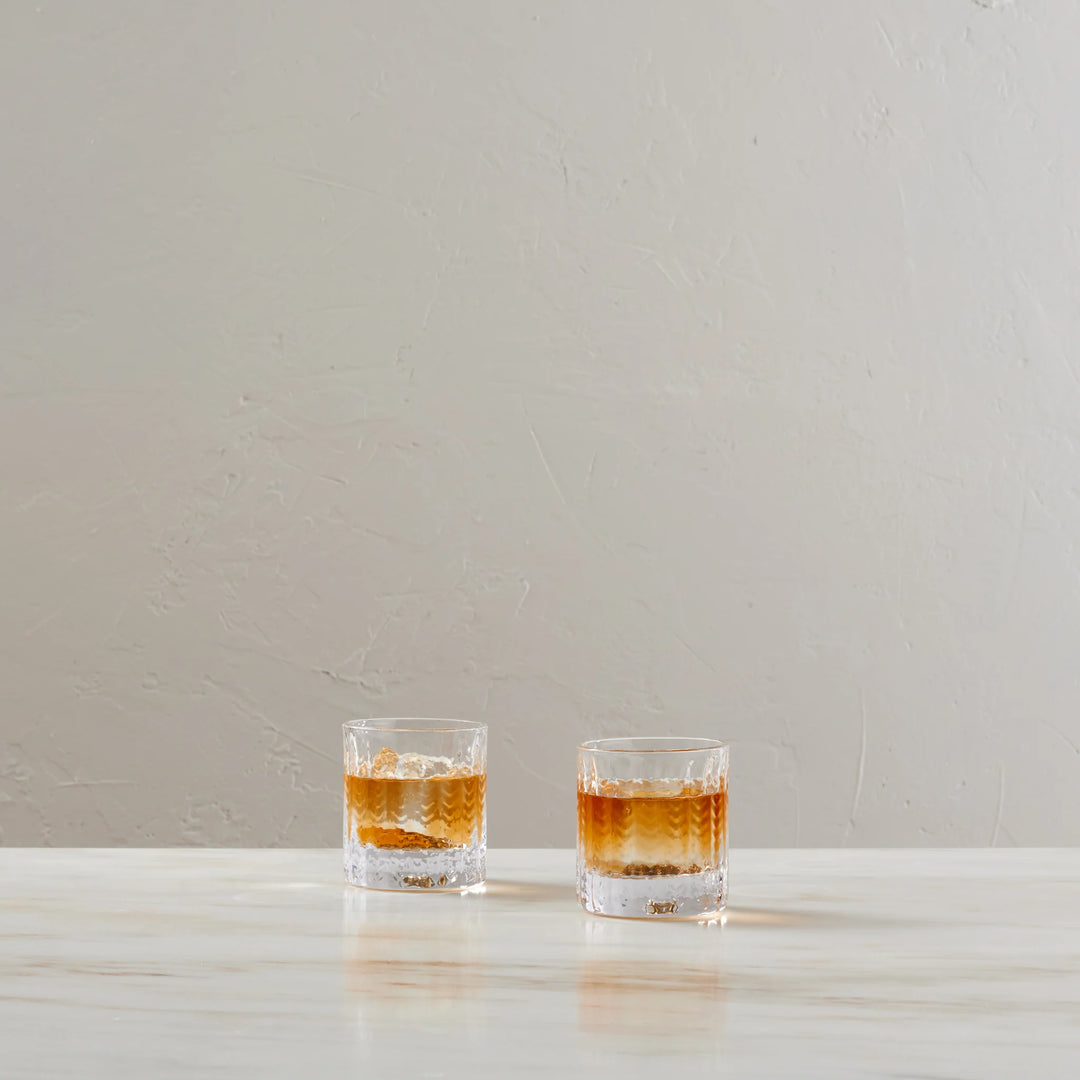 Two clear textured rocks glasses containing an amber liquid, placed on a light marble surface against a neutral beige wall.