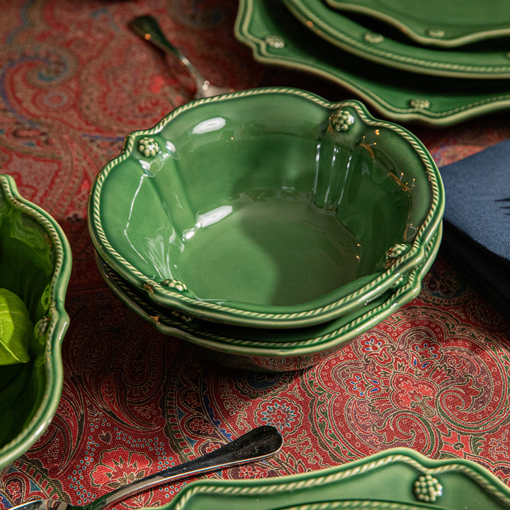A stack of green ceramic bowls with decorative rope-trimmed edges and raised floral details, arranged on a red paisley tablecloth with matching green plates and silver spoons.