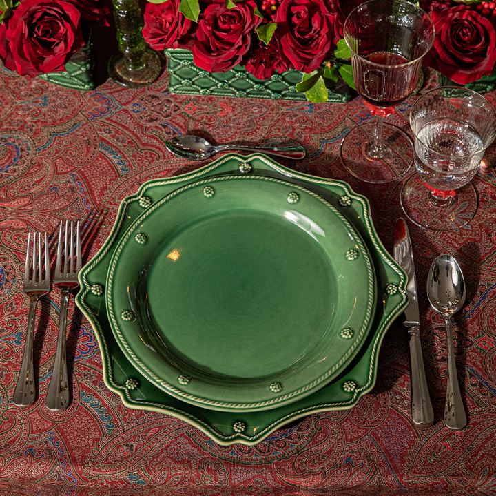 A place setting featuring a green ceramic charger plate with a scalloped rim and decorative beaded edge, layered over a matching green dinner plate. The plates are arranged on a red paisley tablecloth alongside silver flatware, clear glassware, and a red rose centerpiece in the background.