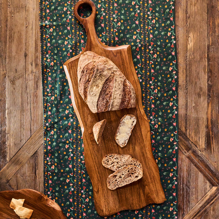 A rustic wooden serving board with a loop handle, styled with sliced artisan bread on top, set on a dark floral runner over a wooden table.