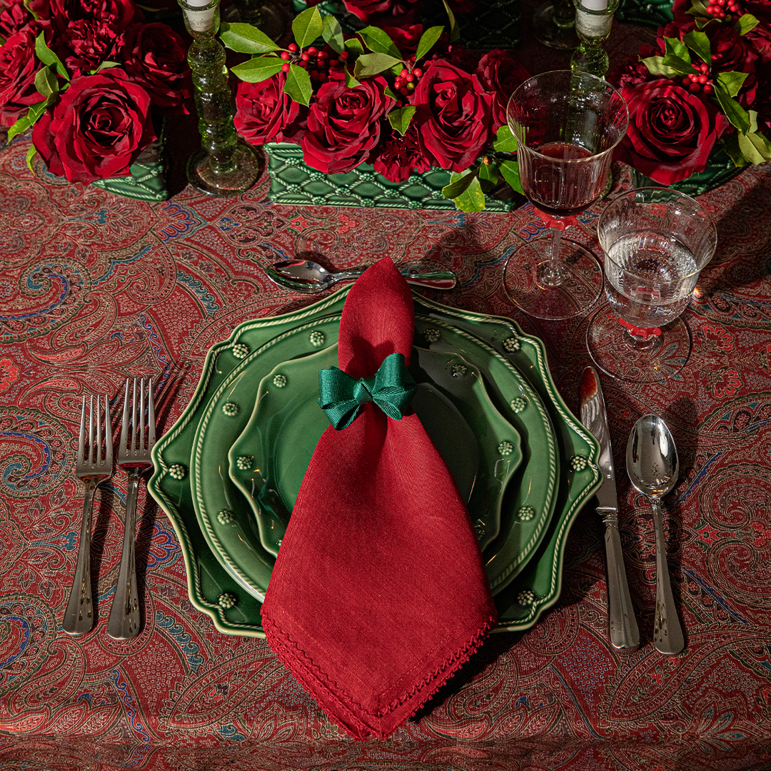 Dining table setting with red napkin, green plates, and red roses on a patterned tablecloth.