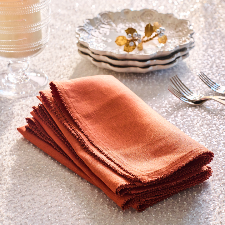 Stack of orange napkins on a textured surface with a candle and stack of decorative white cocktail plates  in the background.