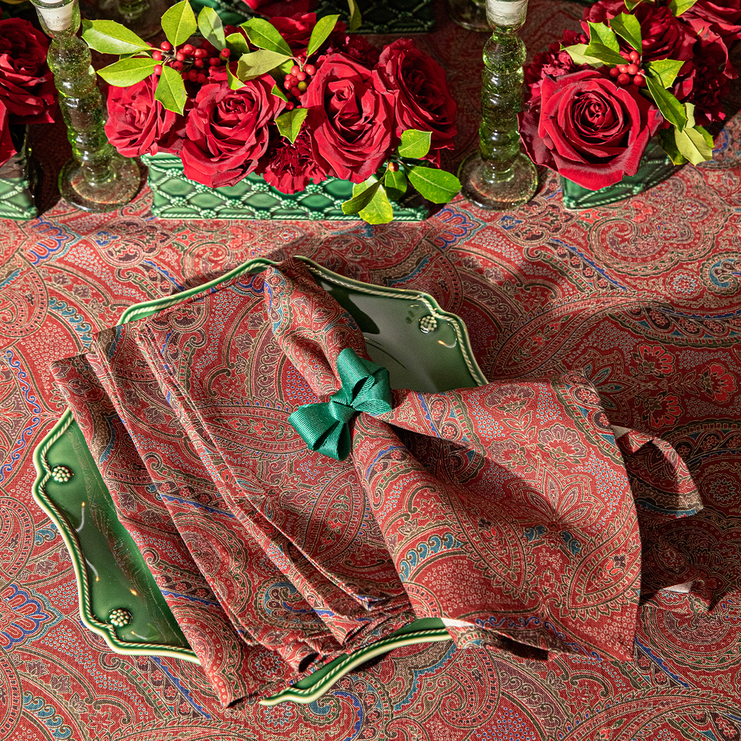 Decorative table setting with red paisley napkins, green napkin holder, and red roses on a patterned tablecloth.