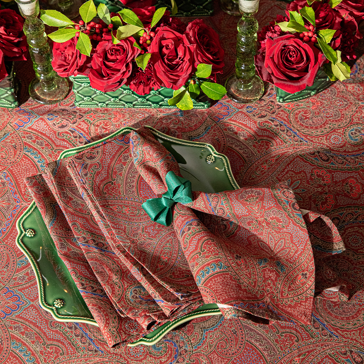Decorative table setting with red paisley napkins, green napkin holder, and red roses on a patterned tablecloth.