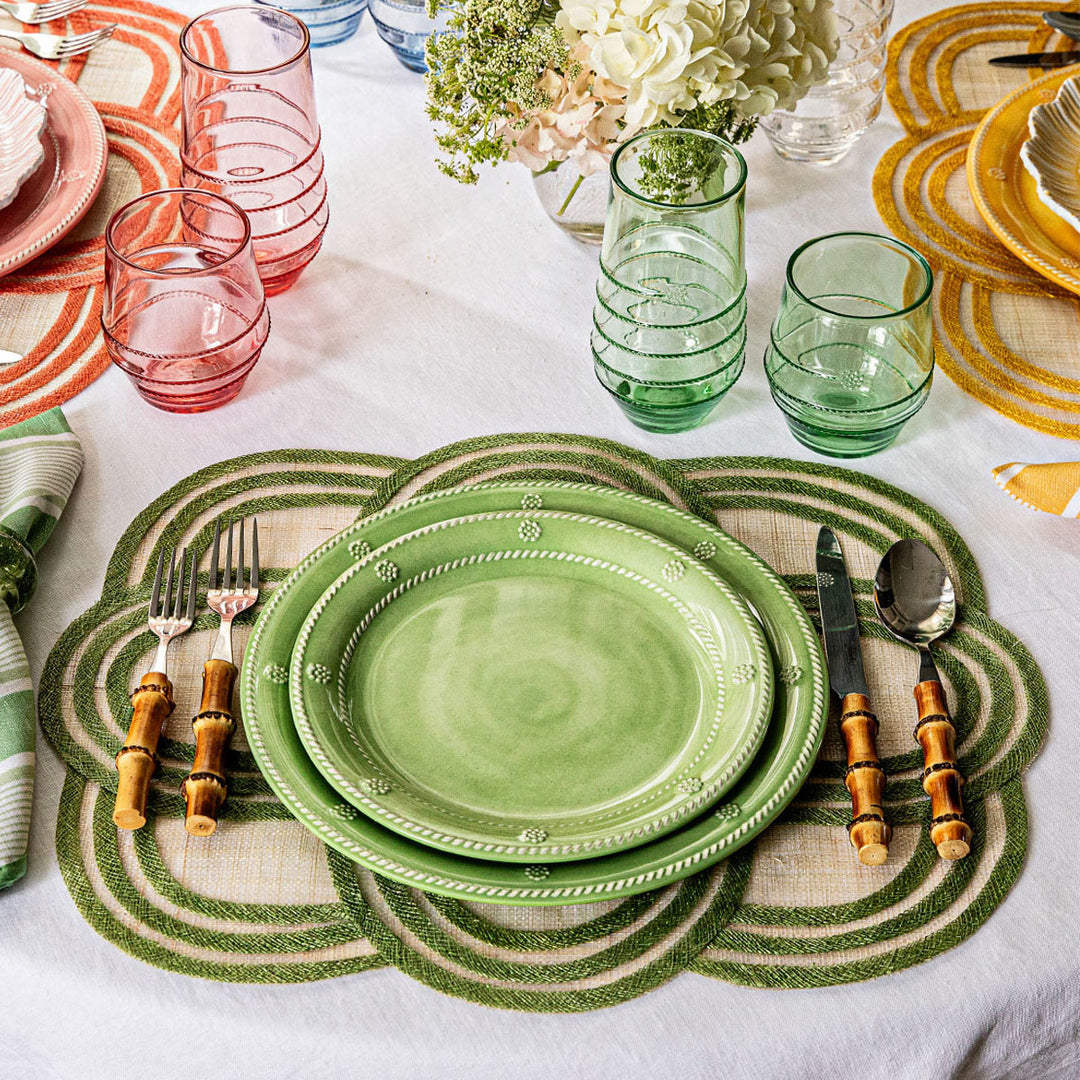 Green ceramic plates with cutlery on a decorative placemat with colorful tableware in the background.