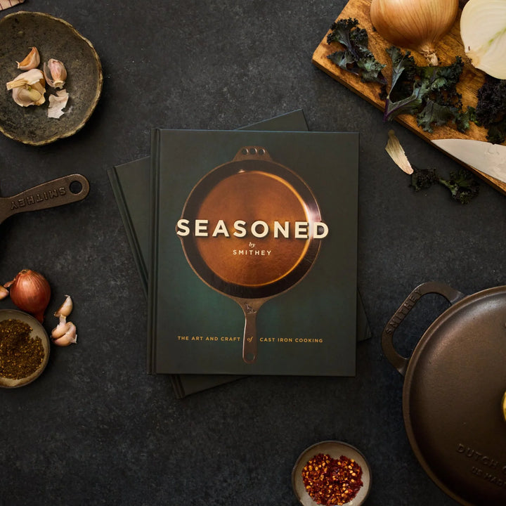 A styled flat-lay of the book 'Seasoned: The Art and Craft of Cast Iron Cooking' by Smithey, featuring a dark green cover with a copper-colored cast iron skillet illustration. The book is surrounded by cooking ingredients and cast iron cookware on a dark textured surface.