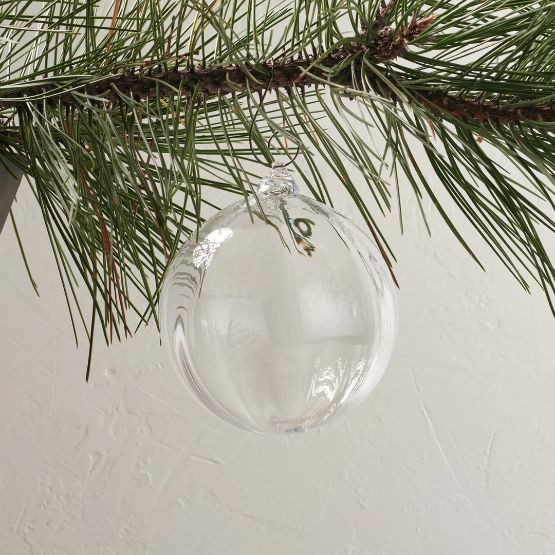 A clear glass Christmas ornament hanging from a pine tree branch against a textured off-white wall.