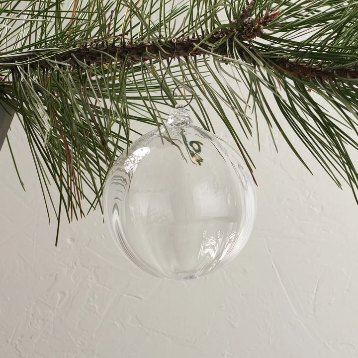 A clear glass Christmas ornament hanging from a pine tree branch against a textured off-white wall.