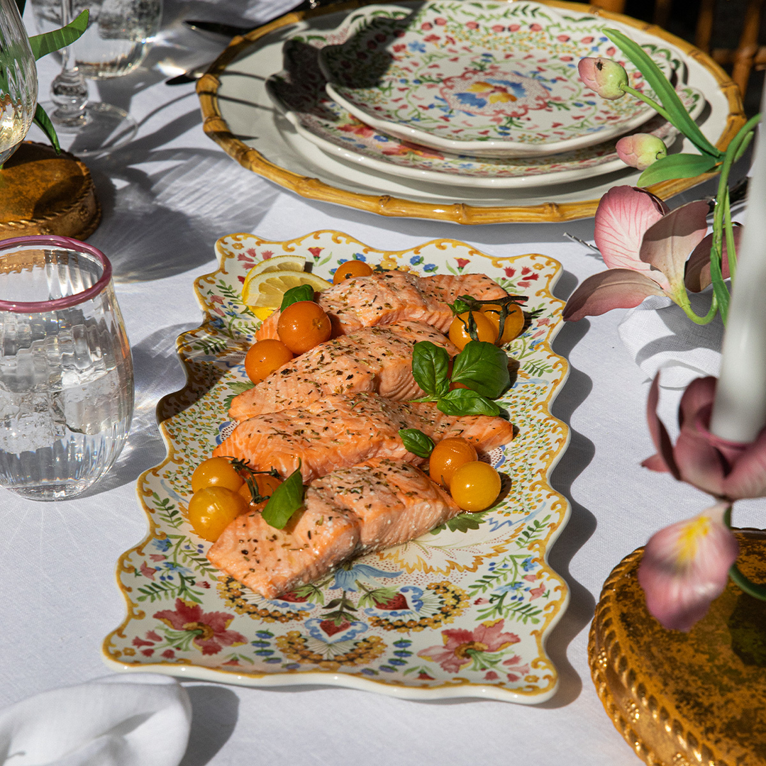 Plated salmon dish on a decorative plate with a floral tablecloth and glasses in the background.