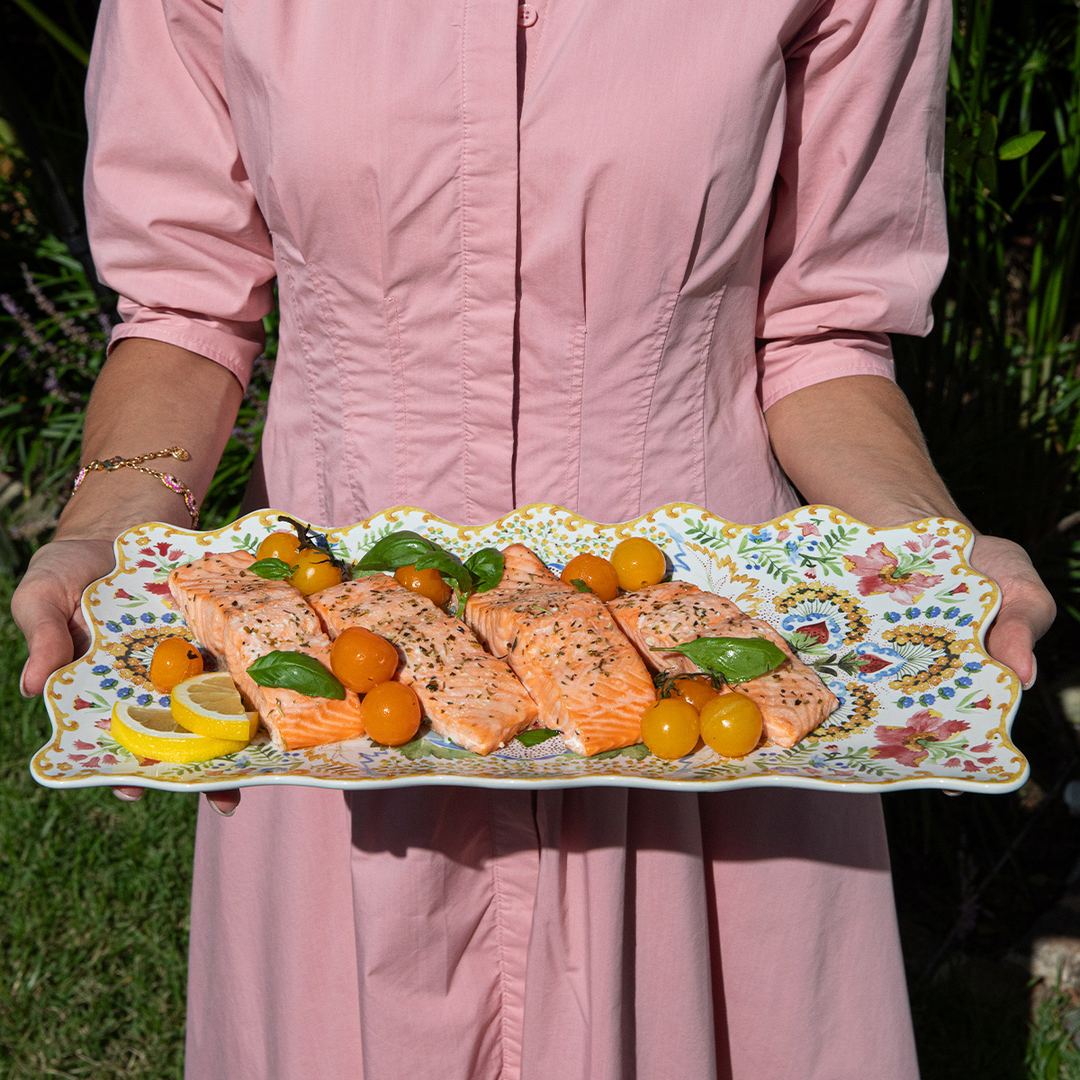 Person holding a decorative plate with salmon and vegetables outdoors.