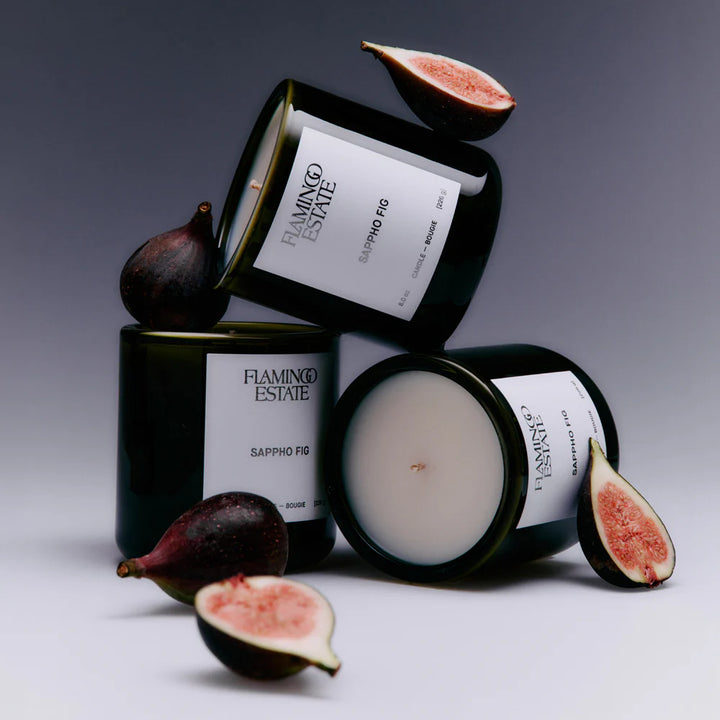Three dark glass jar candles with white labels reading FLAMINGO ESTATE SAPPHO FIG are arranged on a light gray studio background, with whole and sliced figs placed around and on top of the jars.