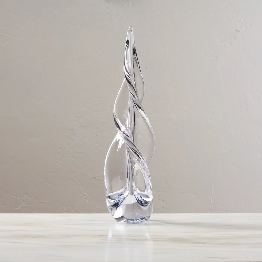 A clear glass christmas tree sculpture with a twisted, flame-like form standing on a light marble surface against a neutral beige wall.