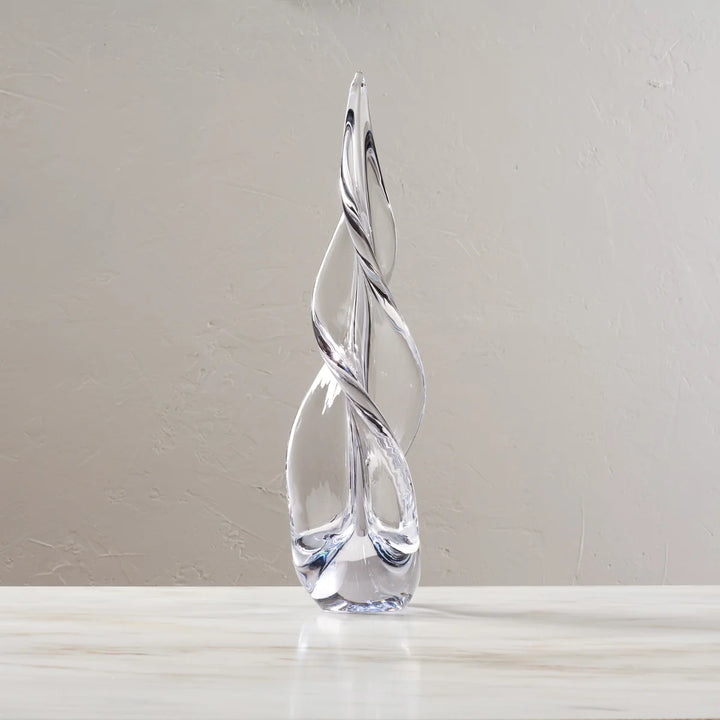 A clear glass christmas tree sculpture with a twisted, flame-like form standing on a light marble surface against a neutral beige wall.