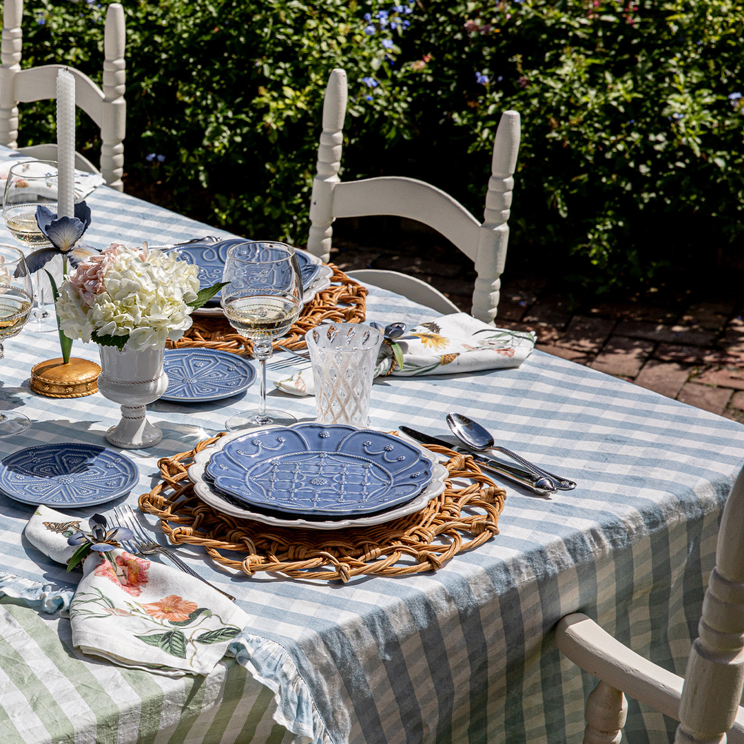 Dining table set with blue plates, glasses, and cutlery on a striped tablecloth outdoors.