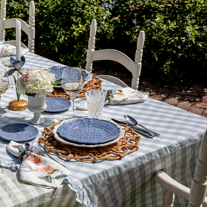 Dining table set with blue plates, glasses, and cutlery on a striped tablecloth outdoors.