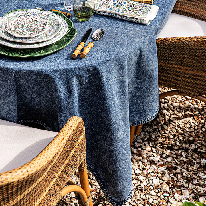 Dining table set with blue tablecloth, plates, and cutlery outdoors on a gravel surface.