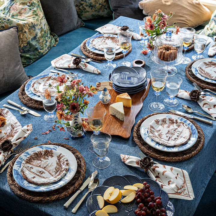 An elegantly set dining table in a cozy living room environment, featuring a blue tablecloth, matching blue and white patterned plates, woven placemats, floral napkins, and glassware.