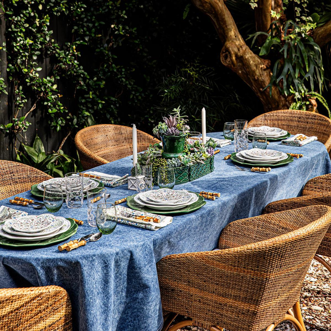 Dining table set for a meal outdoors with a blue tablecloth, green centerpieces, and wicker chairs.