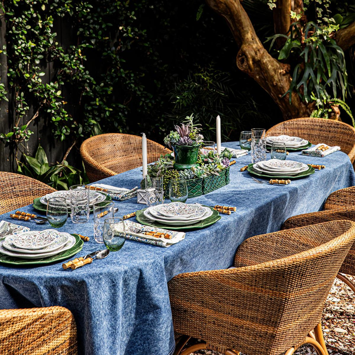Dining table set for a meal outdoors with a blue tablecloth, green centerpieces, and wicker chairs.