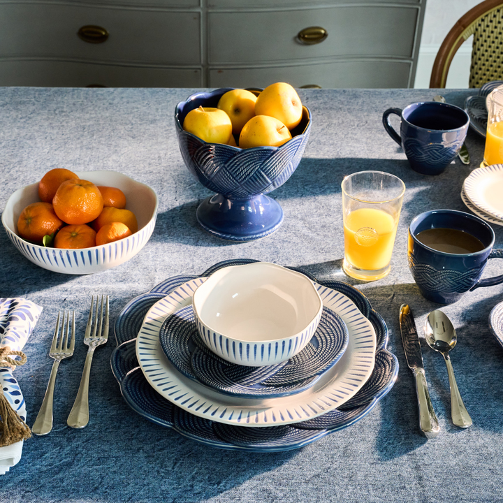 A styled breakfast table setting featuring a coordinated navy blue and white ceramic tableware collection, including plates, bowls, mugs, and a pedestal bowl of fruit, arranged on a blue tablecloth.