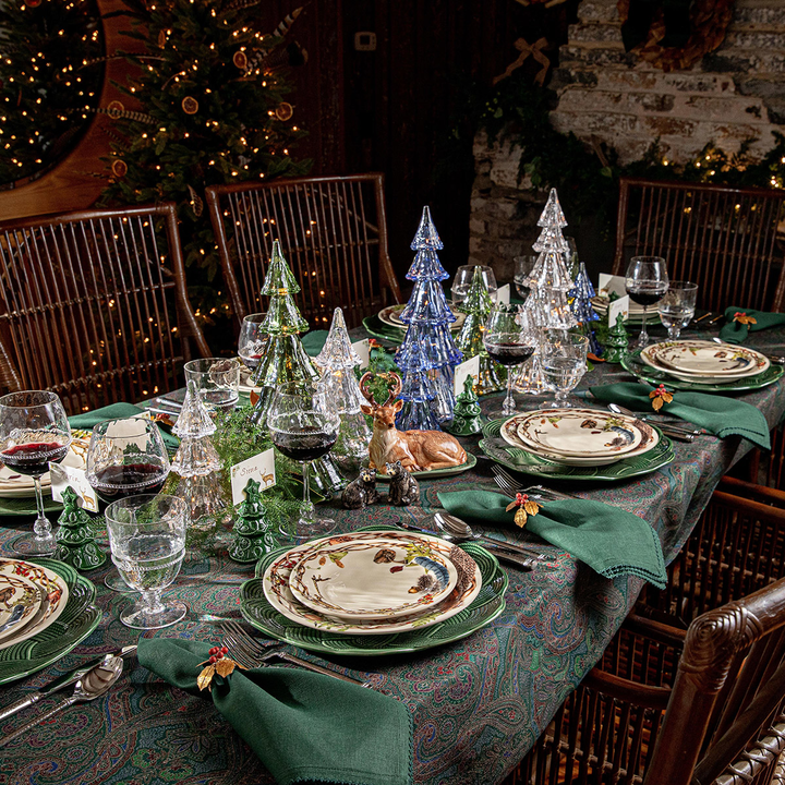 A festive holiday table setting in a rustic dining room, featuring green patterned plates, dark green folded napkins with small floral ring napkins, wine glasses, and clear glass miniature Christmas trees on a patterned tablecloth.
