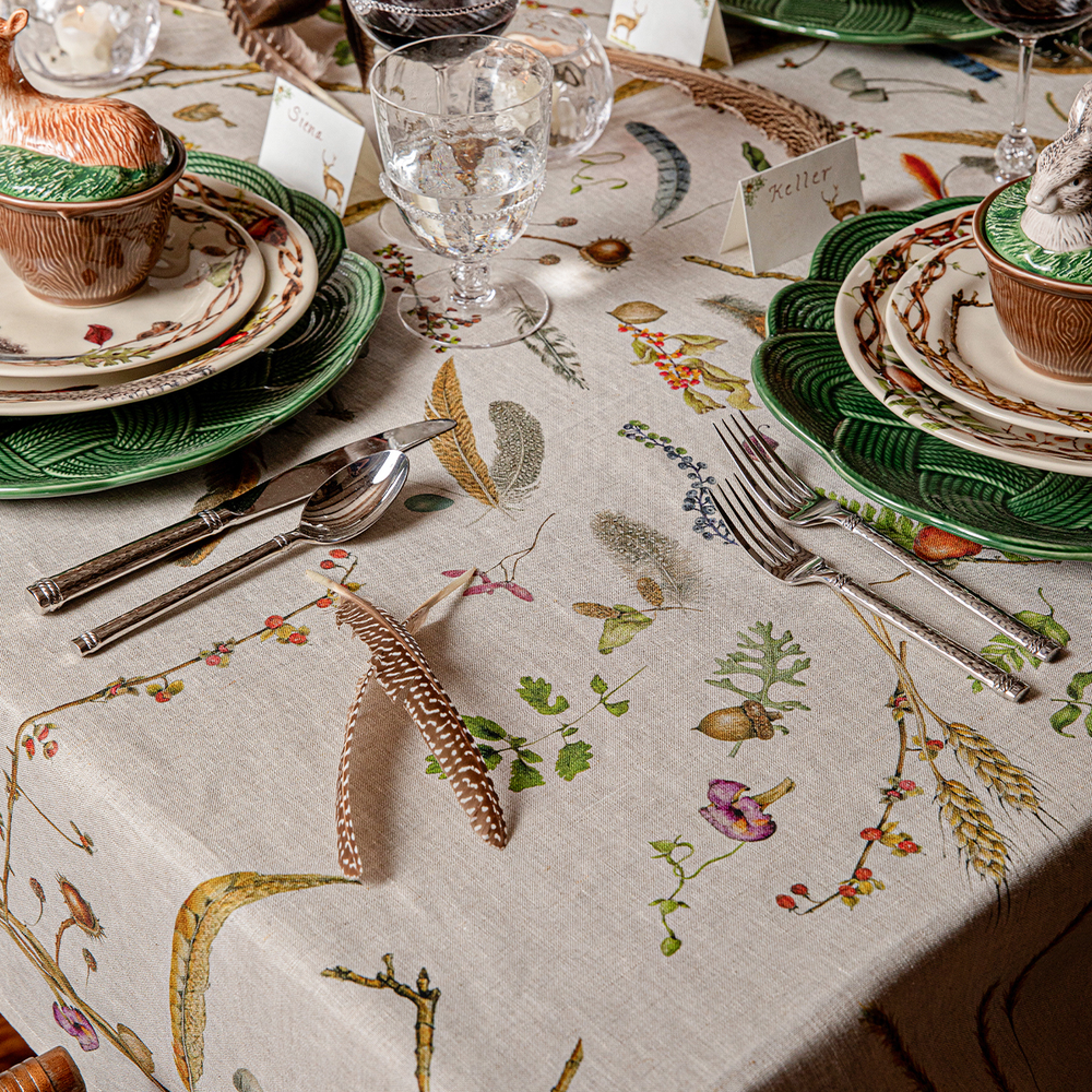 A detailed close-up of a rustic, nature-themed tablecloth featuring a botanical pattern with leaves, acorns, wildflowers, and feathers. The table is styled with matching green and white plates, silver flatware, clear glass goblets, and small decorative ceramic pots.