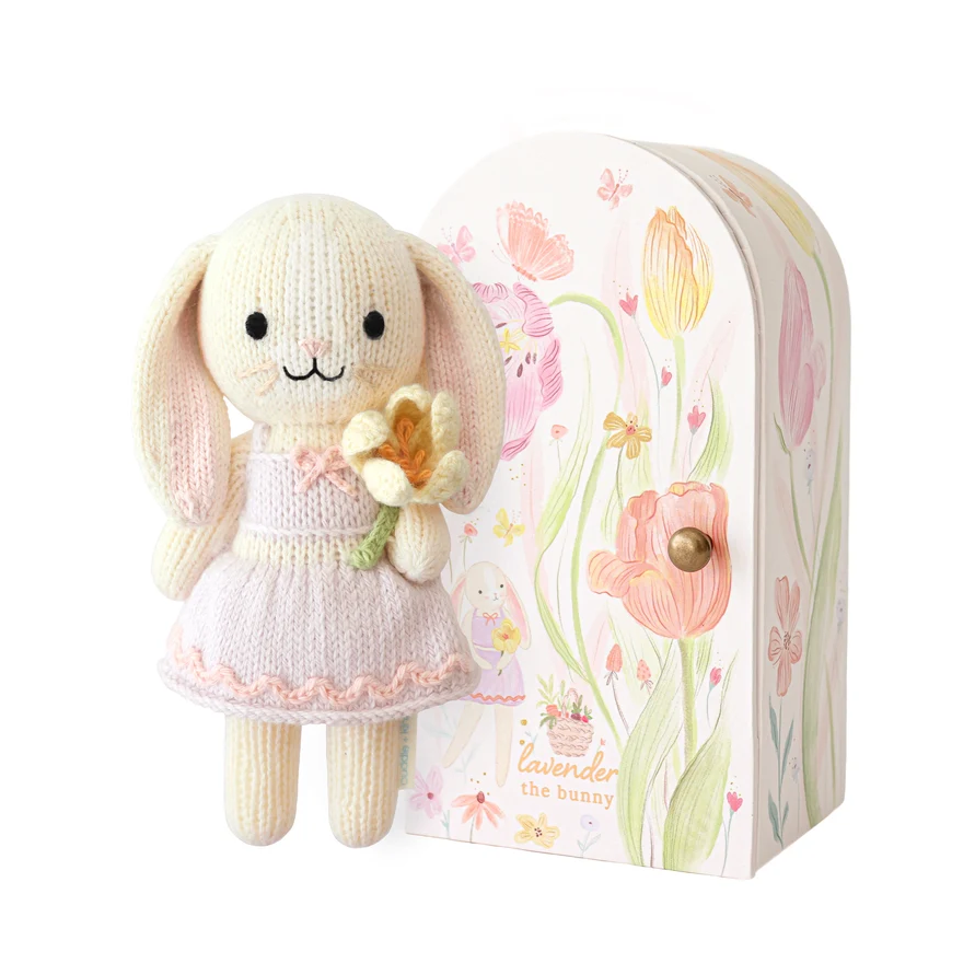 Knitted bunny toy with a pink dress holding flowers, standing next to a floral-patterned box
