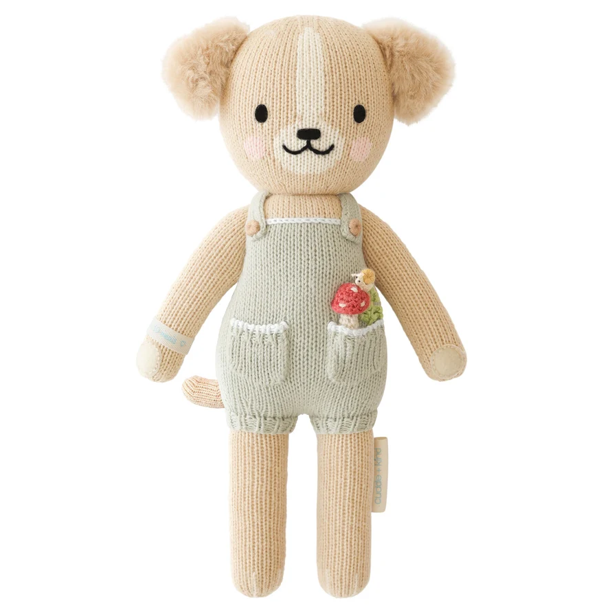 A knitted puppy plush toy wearing light sage green overalls with a small mushroom in his pocket