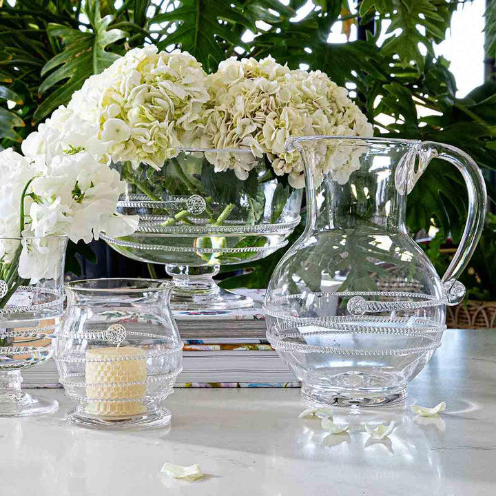 Clear glass pitcher and candle on a table with white flowers and books in the background