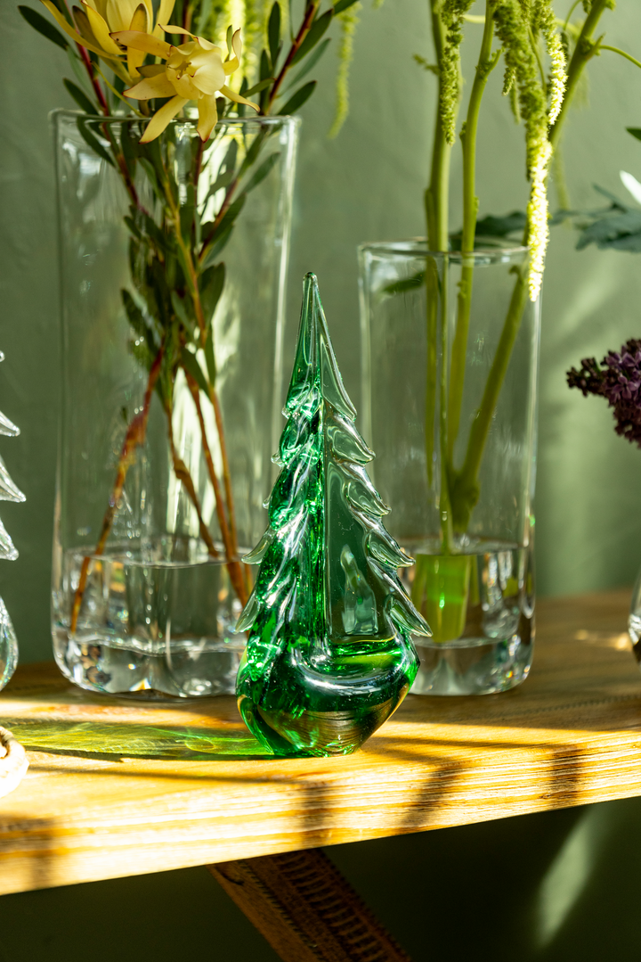 Green glass Christmas tree ornament on a wooden mantle with clear glass vases containing plants.