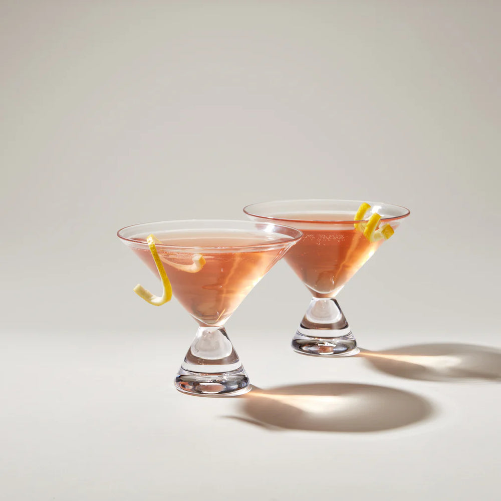 Two pink cocktails with lemon twists in Simon Pearce martini glasses on a light background