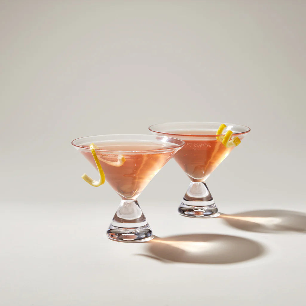 Two pink cocktails with lemon twists in Simon Pearce martini glasses on a light background