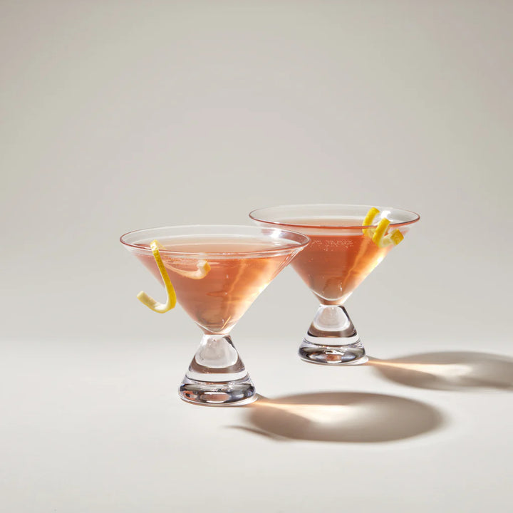 Two pink cocktails with lemon twists in Simon Pearce martini glasses on a light background