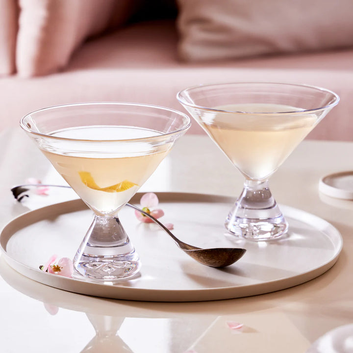 Two Simon pearce martini glasses with lemon drop martinis on a white plate with a spoon and cherry blossoms, against a soft pink background.