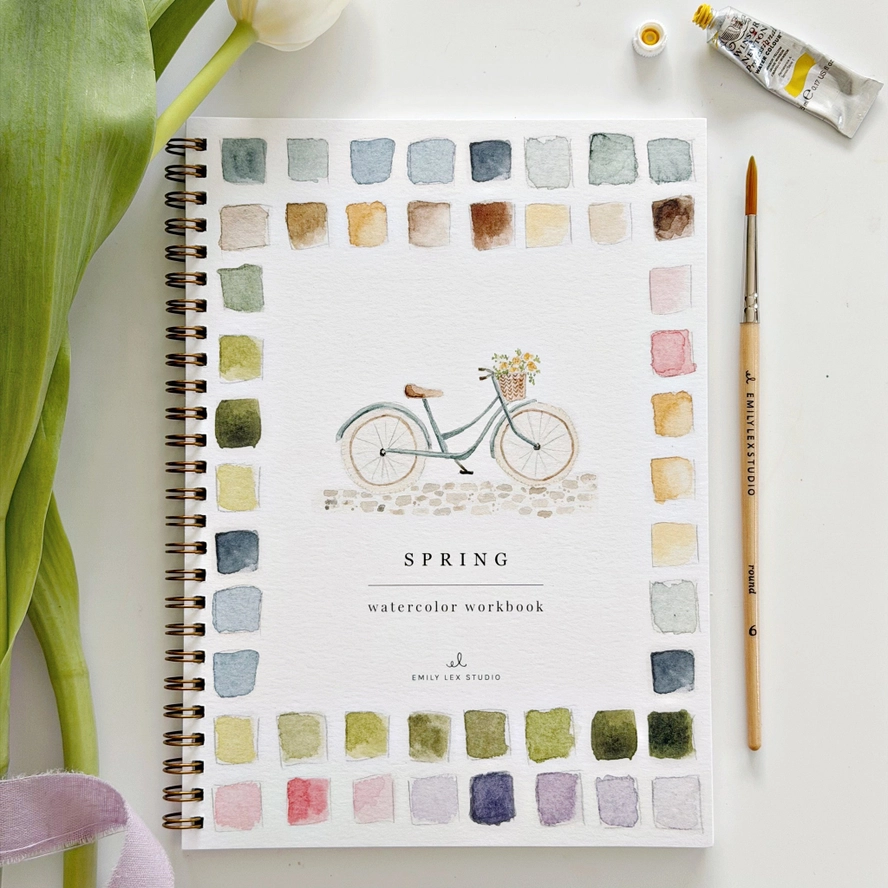 Spring watercolor workbook with a bicycle illustration and color swatches, surrounded by art supplies 