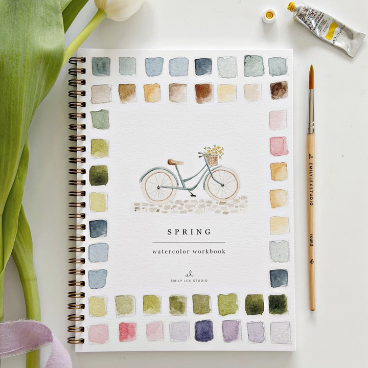 Spring watercolor workbook with a bicycle illustration and color swatches, surrounded by art supplies 