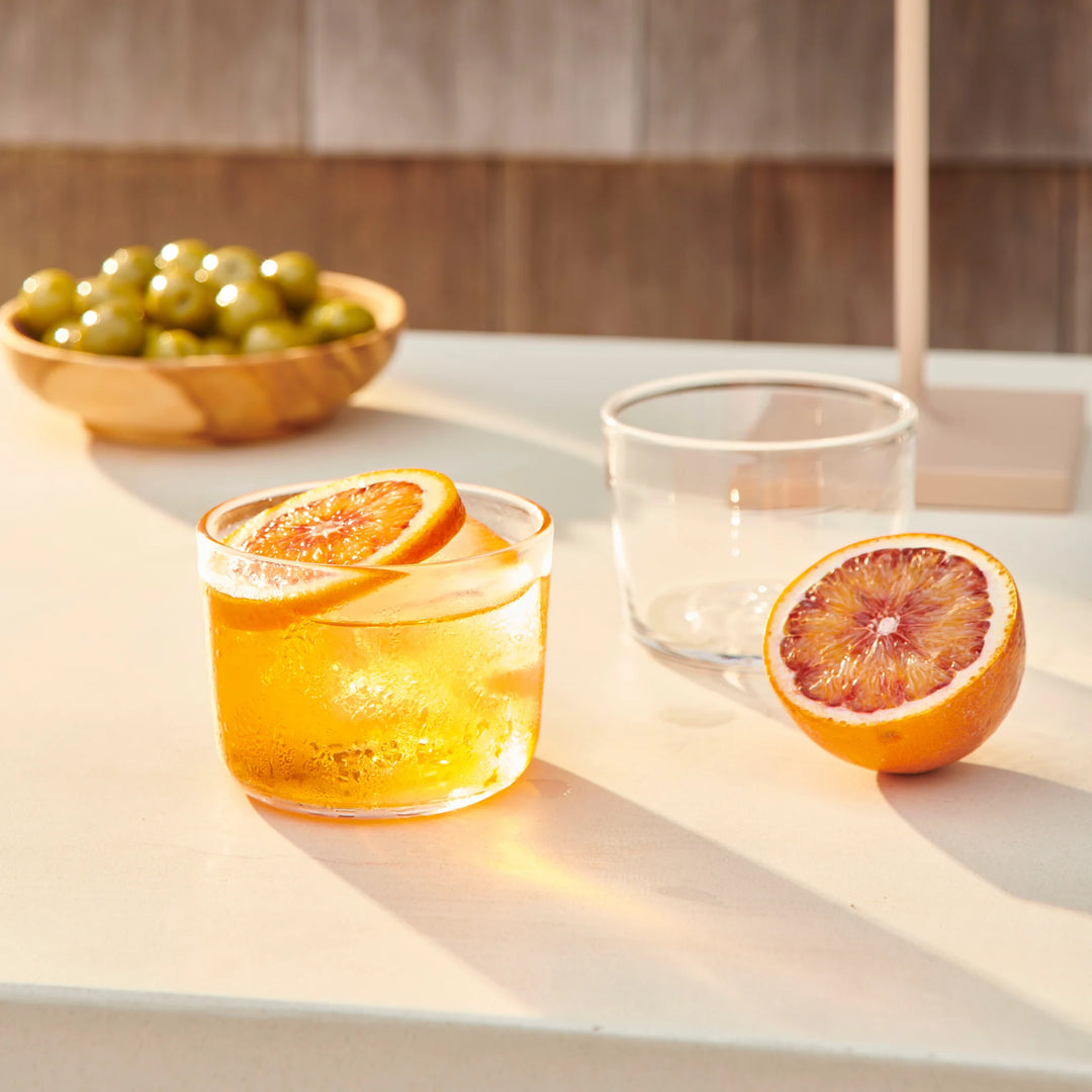 Glass of orange juice with a slice of orange on a light surface, next to a whole orange and a bowl of green olives.