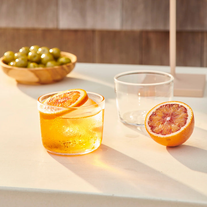 Glass of orange juice with a slice of orange on a light surface, next to a whole orange and a bowl of green olives.