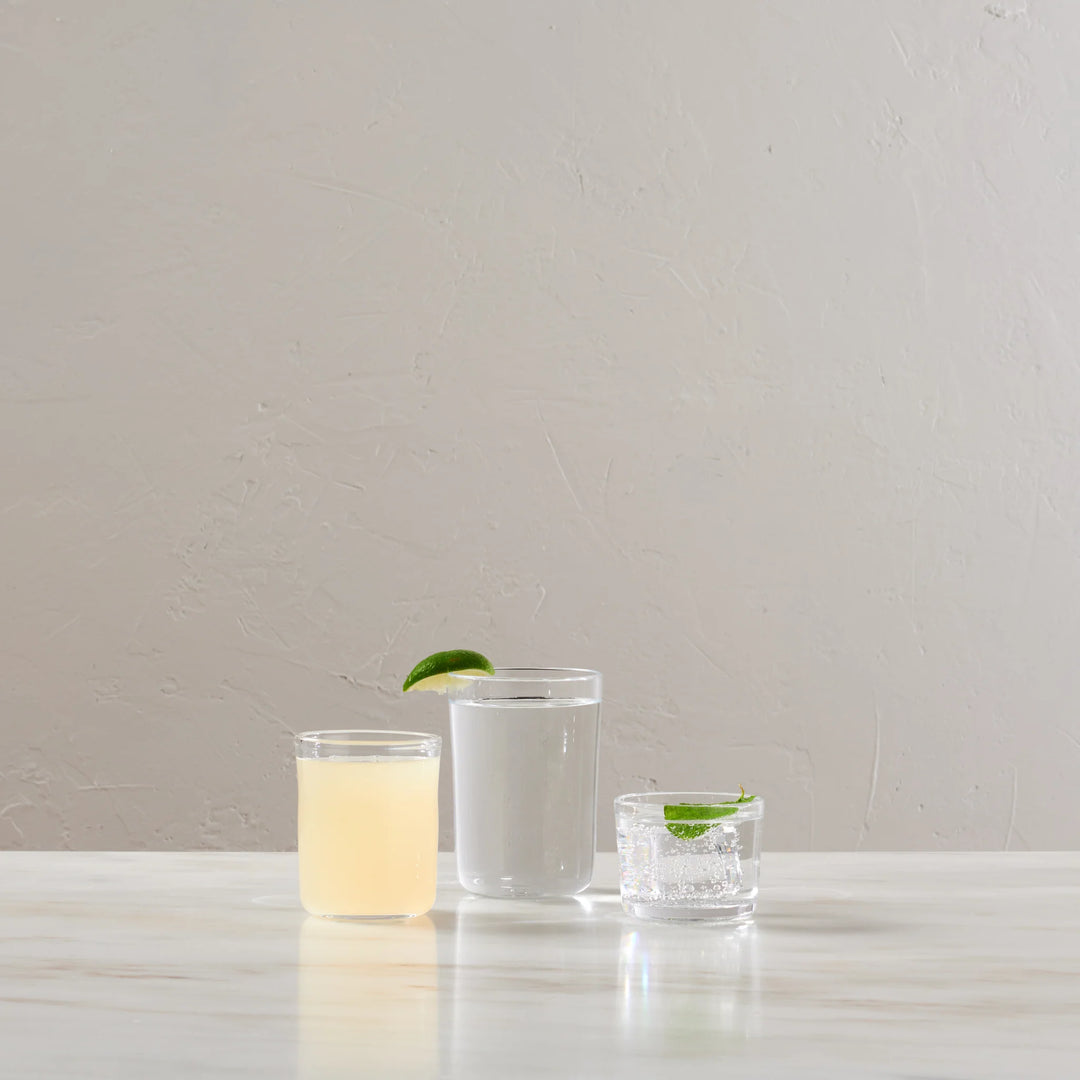 Three clear glass tumblers with different designs and drink contents on a light marble surface against a textured beige wall.