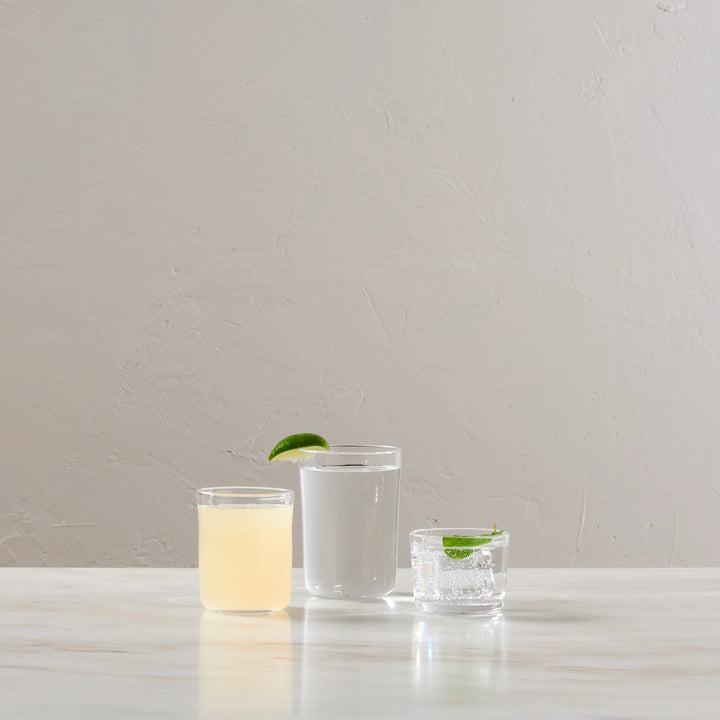 Three clear glass tumblers with different designs and drink contents on a light marble surface against a textured beige wall.