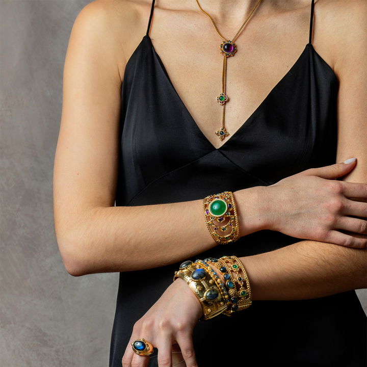 A close-up of a model wearing an ornate gold jewelry set, including a long drop necklace, a wide cuff bracelet, a stack of bangles, and a ring, all decorated with green and blue stones, paired with a black satin dress against a gray background.