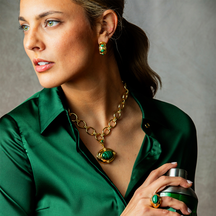 A woman in a green satin shirt models a matching green and gold jewelry set, including a necklace, earrings, and ring, while holding a green cocktail shaker against a textured gray background.