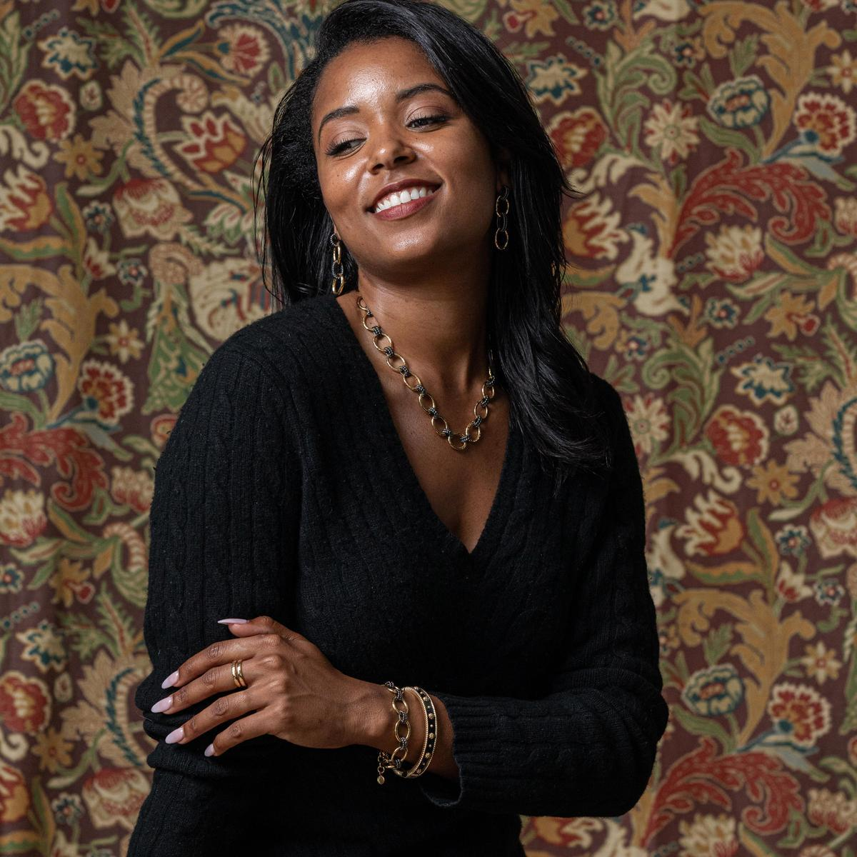Woman wearing a black sweater and gold jewelry with a decorative patterned wall behind her