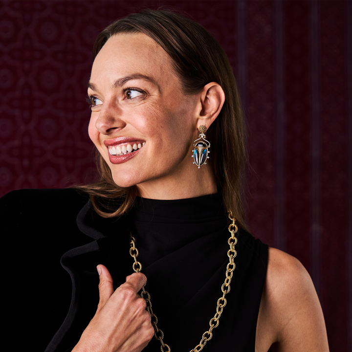 Woman wearing gold scarab earrings and a necklace against a patterned red background
