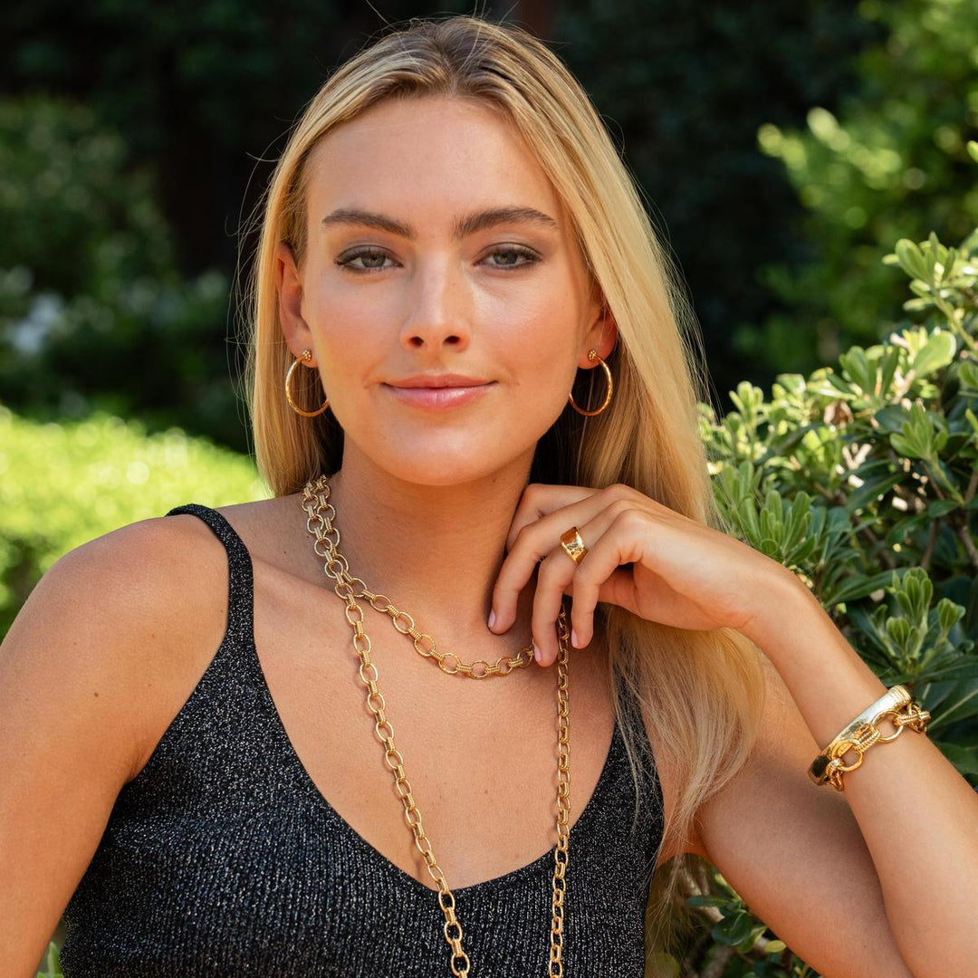 Woman wearing gold jewelry outdoors with greenery in the background