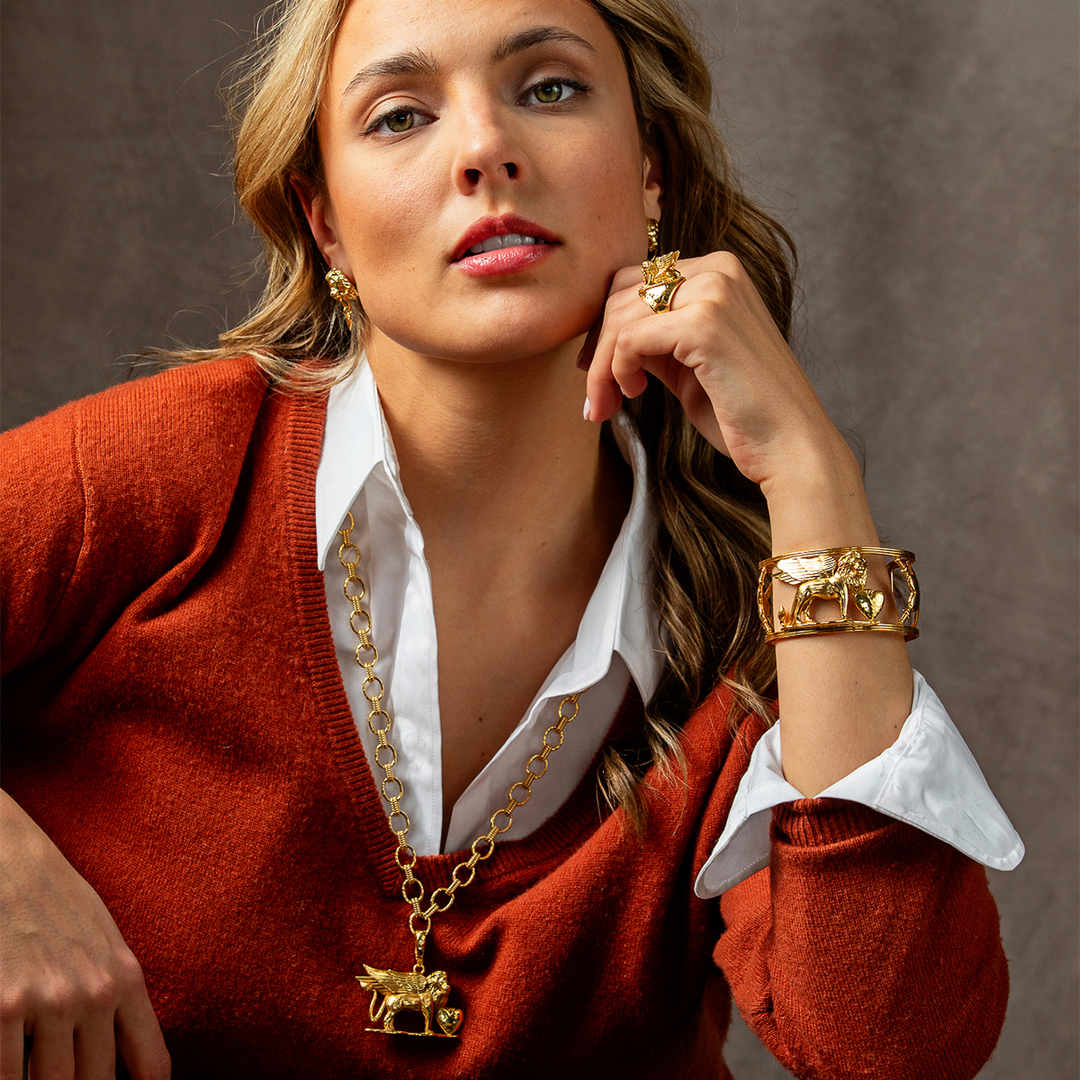 A close-up lifestyle shot of a woman wearing a gold animal-themed jewelry set, including a necklace with a lion pendant, a matching lion ring, and a lion cuff bracelet, styled over a rust-colored sweater and white collared shirt against a textured brown background.
