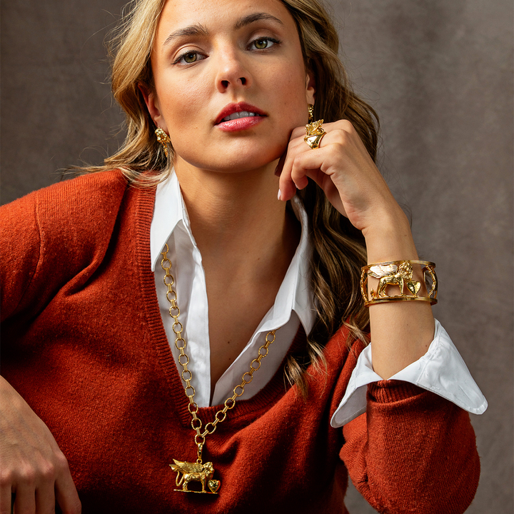A close-up lifestyle shot of a woman wearing a gold animal-themed jewelry set, including a necklace with a lion pendant, a matching lion ring, and a lion cuff bracelet, styled over a rust-colored sweater and white collared shirt against a textured brown background.