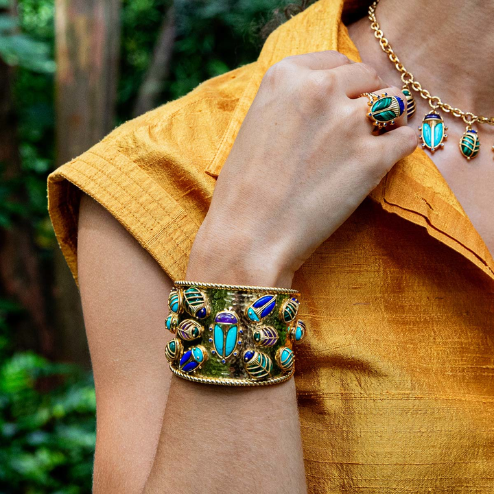 Close-up of a person wearing gold jewelry with beetles made of colorful stones against a blurred natural background