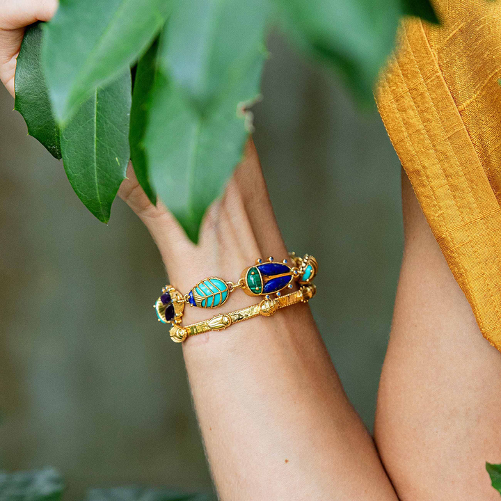 Gold bracelet with green and blue beetle shaped stones on a person's wrist, held against a leafy background.