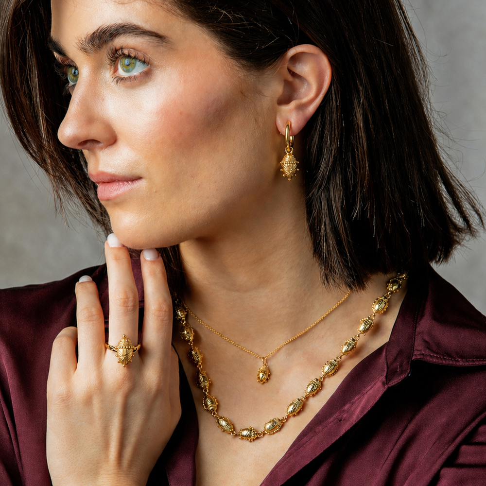 A close-up lifestyle shot of a woman wearing a gold jewelry set, including a textured drop earring, a matching necklace, and a ring, paired with a deep burgundy satin shirt against a neutral gray background.
